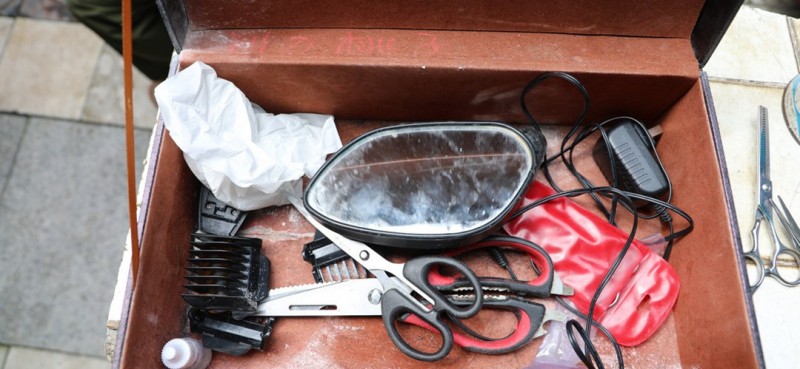 一把电推剪子、一把普通梳子、一块用废旧雨伞布制成的围布、一个电动车废旧后视镜、一个由红酒箱做成的工具箱，这就是邹勤敏用来剪发的装备。